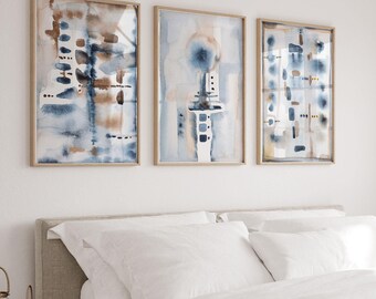 May include: A white bed with a beige knitted throw blanket and three framed abstract watercolour paintings above the headboard. The paintings are in shades of blue, brown, and white.