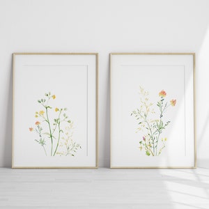 May include: Two framed watercolor prints of wildflowers. The print on the left features yellow wildflowers with green stems. The print on the right features orange wildflowers with green stems.