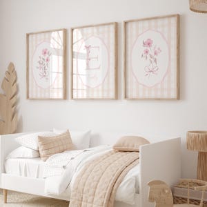 May include: Three framed floral art prints with a pink and beige color scheme hang above a white bed. The prints feature pink flowers and the letter "E" with the name "Emily". The bed has a beige quilt and pillows.