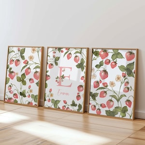 Op de afbeelding: Drie ingelijste prints met een witte achtergrond en een patroon van roze aardbeien en groene bladeren. De middelste print heeft de letter "E" in roze met de naam "Emma" eronder.