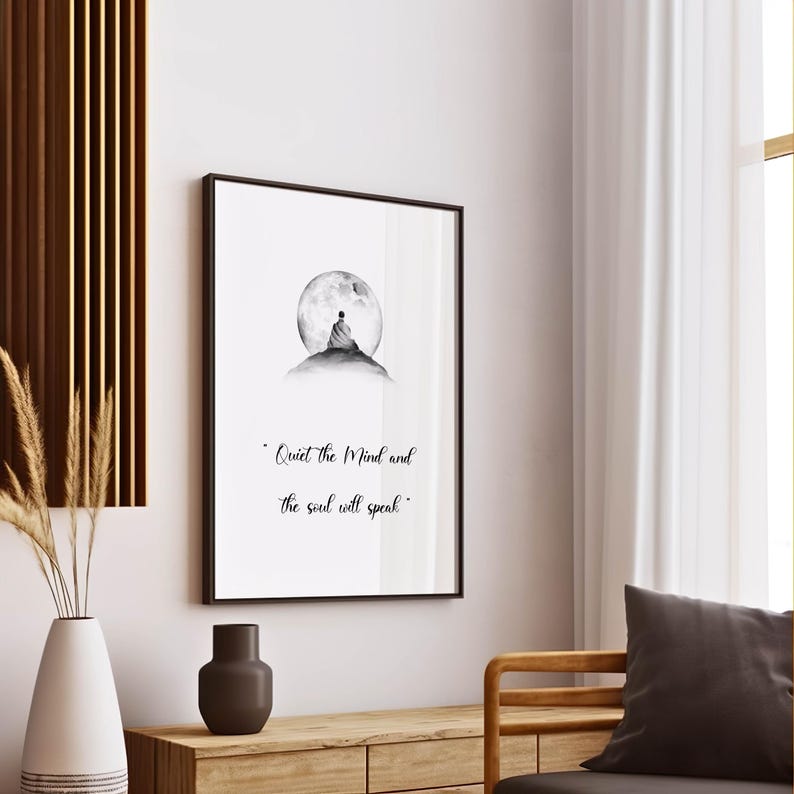 May include: A framed black and white print featuring a person meditating on a hill in front of a full moon. The text "Quiet the Mind and the soul will speak" is below the image. The print is displayed on a white wall.