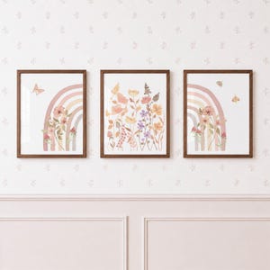 May include: A set of three framed wall art prints with dark wood frames. The outer prints feature a muted rainbow with floral accents and butterflies. The centre print displays a collection of wildflowers in shades of yellow, pink, and purple.