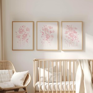May include: Three framed floral art prints with pink watercolour flower designs. The prints are in gold frames and hang above a wooden cot. A wicker chair with white cushions is in the foreground.