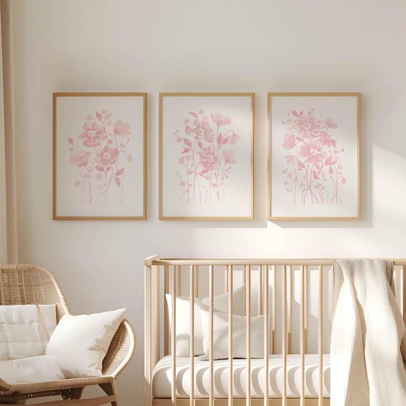 May include: Three framed floral art prints with pink watercolour flower designs. The prints are in gold frames and hang above a wooden cot. A wicker chair with white cushions is in the foreground.
