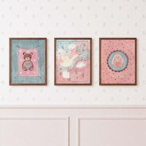May include: Three rectangular wall art prints in dark wood frames. The left print shows a brown bear on a pink and blue striped background. The center print features a yellow goose in a pink sky. The right print displays a pink rabbit in a circle.