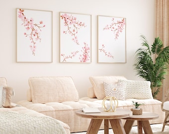 以下が含まれることがあります： 白い背景にピンクの桜の花が描かれた水彩画が3枚、額装されています。絵画は、薄いベージュのソファの上に2つのクッションを置いた白い壁に飾られています。ソファの前に木製のコ​​ーヒーテーブルが置かれています。