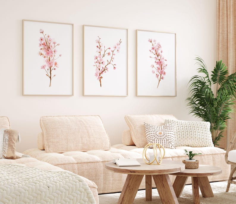 May include: Three framed watercolor paintings of pink cherry blossoms on a white background. The paintings are hung on a white wall above a light beige sofa with pillows.