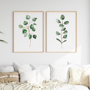 May include: Two framed watercolor prints of eucalyptus branches with green leaves on a white background. The prints are hanging on a white wall above a white couch with pillows.