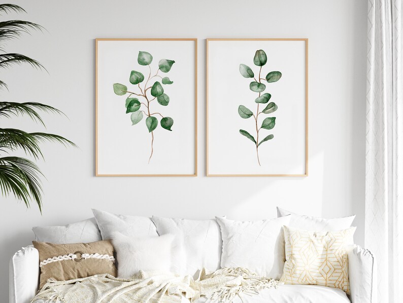 May include: Two framed watercolor prints of eucalyptus branches with green leaves on a white background. The prints are hanging on a white wall above a white couch with pillows.