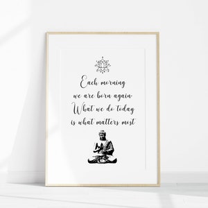 May include: A black and white printable wall art print with a quote that reads "Each morning we are born again. What we do today is what matters most." The quote is centered on a white background with a black lotus flower design at the top and a black silhouette of a meditating Buddha at the bottom.