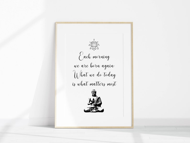 May include: A black and white printable wall art print with a quote that reads "Each morning we are born again. What we do today is what matters most." The quote is centered on a white background with a black lotus flower design at the top and a black silhouette of a meditating Buddha at the bottom.