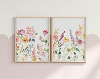 May include: Two vertical rectangular wall art prints in light wood frames, each featuring a watercolor illustration of a wildflower meadow with pink, yellow, and purple blossoms on thin green stems against a plain white background.
