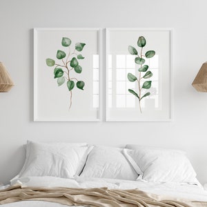 May include: Two white framed prints of watercolor paintings of green eucalyptus branches on a white wall above a bed with white bedding and a tan throw blanket.