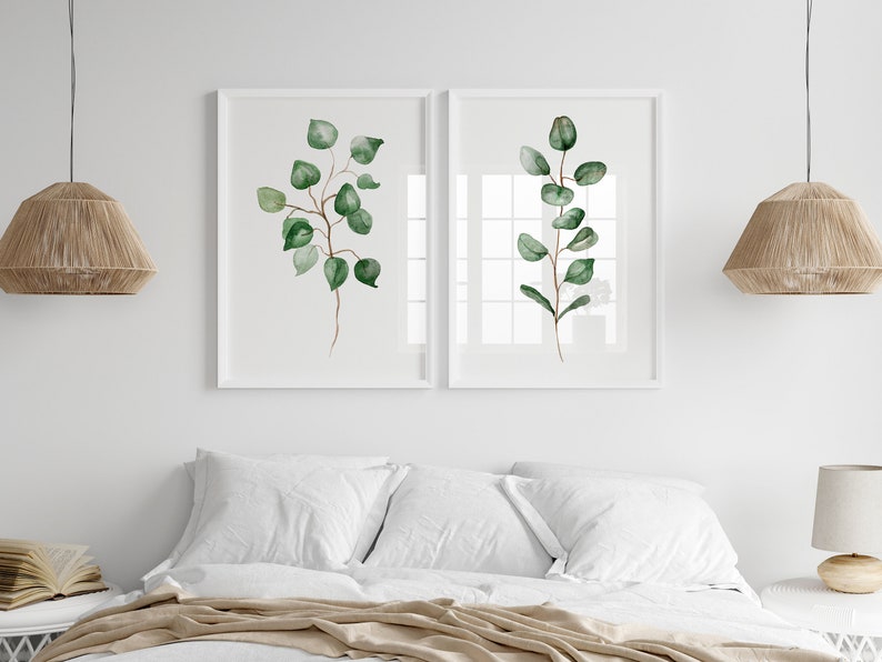 May include: Two white framed prints of watercolor paintings of green eucalyptus branches on a white wall above a bed with white bedding and a tan throw blanket.
