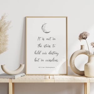 Puede incluir: Un cuadro enmarcado con borde dorado que presenta una cita de William Shakespeare: "No está en las estrellas mantener nuestro destino, sino en nosotros mismos". La obra de arte incluye una ilustración de una luna creciente.