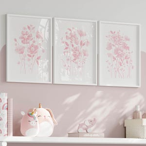 May include: Three framed floral art prints in white frames. The prints depict pink watercolour flowers on a white background. The prints are displayed above a shelf with pink plush toys and decorative items.