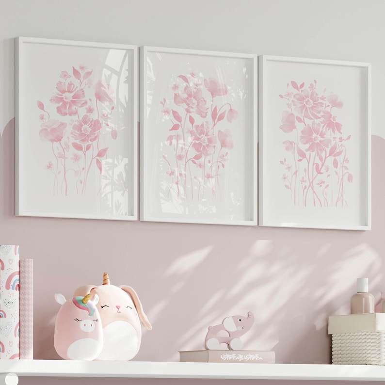May include: Three framed floral art prints in white frames. The prints depict pink watercolour flowers on a white background. The prints are displayed above a shelf with pink plush toys and decorative items.
