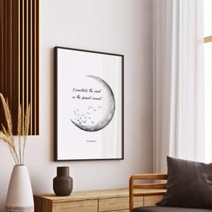 May include: A framed black and white print with the text "Concentrate the mind in the present moment" and a crescent moon design with flying birds. The print is displayed on a wooden table with a vase and decorative items.