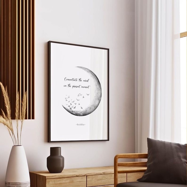 May include: A framed black and white print with the text "Concentrate the mind in the present moment" and a crescent moon design with flying birds. The print is displayed on a wooden table with a vase and decorative items.