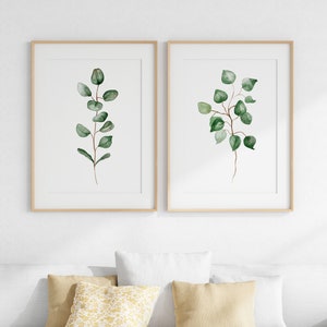 May include: Two framed watercolor prints of green eucalyptus branches on a white background. The prints are in simple wooden frames and are hung on a white wall.