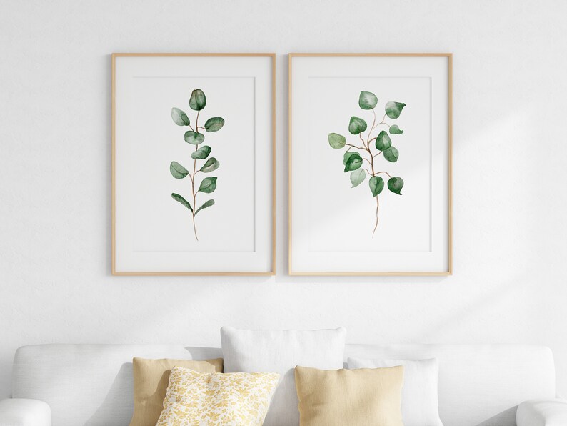 May include: Two framed watercolor prints of green eucalyptus branches on a white background. The prints are in simple wooden frames and are hung on a white wall.