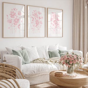 May include: A living room with a white sofa and cushions in white and green. Three framed floral prints in soft pink tones are on the wall. A wooden coffee table holds a bouquet of pink flowers. The room has a light, airy feel.