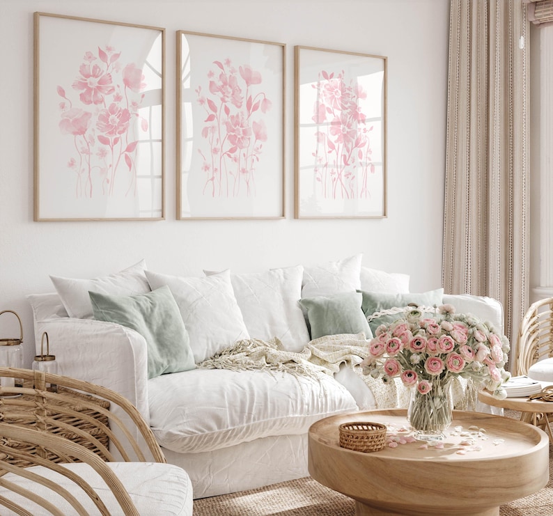 May include: A living room with a white sofa and cushions in white and green. Three framed floral prints in soft pink tones are on the wall. A wooden coffee table holds a bouquet of pink flowers. The room has a light, airy feel.