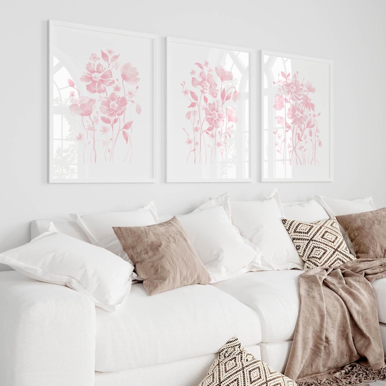 May include: Three framed floral art prints featuring pink watercolour flowers. The prints are above a white sofa with white and beige cushions and a brown throw. The room has a bright, airy feel.