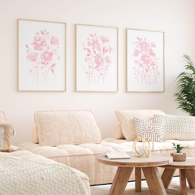 May include: Three framed floral art prints featuring pink watercolor flowers on a white background. The prints are displayed above a light pink sectional sofa with matching cushions. Two round wooden coffee tables and a potted plant complete the room.