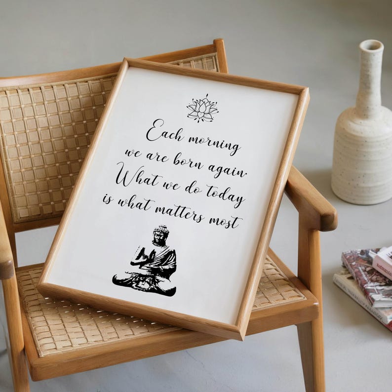 May include: A framed print with a wooden frame, featuring the text "Each morning we are born again. What we do today is what matters most." A black illustration of Buddha is at the bottom. The print is on a wooden chair.