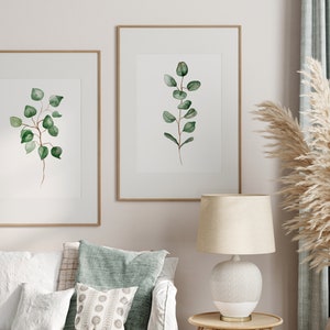 May include: Two framed watercolor prints of green eucalyptus branches on a white background, hanging on a white wall above a white couch with green throw pillows. A white lamp with a beige lampshade sits on a wooden table next to a vase with dried pampas grass.