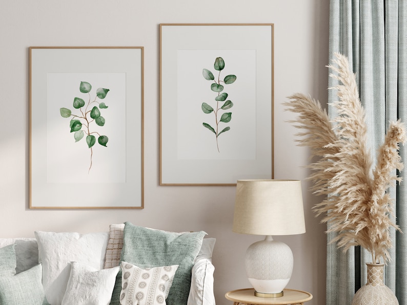 May include: Two framed watercolor prints of green eucalyptus branches on a white background, hanging on a white wall above a white couch with green throw pillows. A white lamp with a beige lampshade sits on a wooden table next to a vase with dried pampas grass.