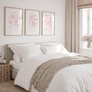 May include: A bedroom with a bed dressed in white linen, a beige knitted throw, and three framed pink floral prints. A wicker bedside table and a vase of pink flowers complete the room's decor.