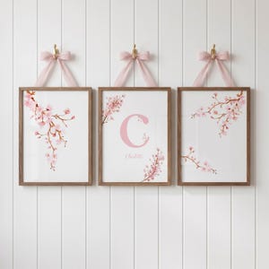 May include: Three rectangular wall art prints in wooden frames, each suspended by a pink ribbon. The prints feature watercolour cherry blossom branches. The centre print displays a large pink letter C and the name Charlotte in script.