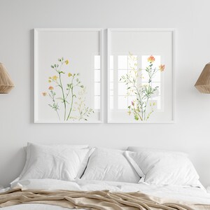 May include: Two framed watercolor prints of delicate wildflowers in shades of yellow, orange, and green. The prints are hung on a white wall above a bed with white bedding and a tan throw.