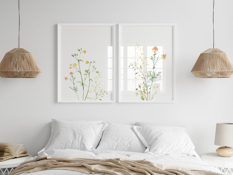 May include: Two framed watercolor prints of delicate wildflowers in shades of yellow, orange, and green. The prints are hung on a white wall above a bed with white bedding and a tan throw.