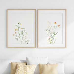 May include: Two framed watercolor prints of delicate wildflowers with green stems and yellow and orange flowers. The prints are in simple wooden frames and are hung on a white wall.