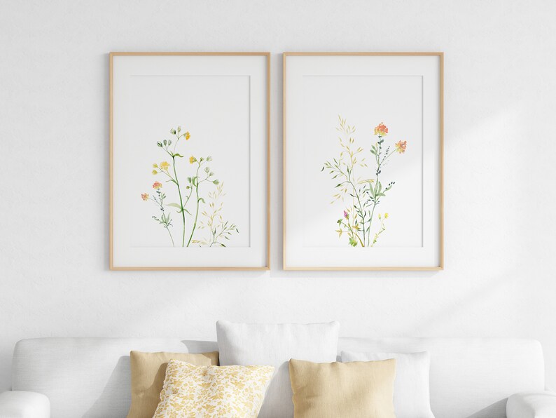 May include: Two framed watercolor prints of delicate wildflowers with green stems and yellow and orange flowers. The prints are in simple wooden frames and are hung on a white wall.