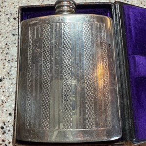 May include: A silver-colored flask with a textured surface and a screw-on cap. The flask is rectangular and has a decorative pattern of vertical lines and diamond shapes. It is housed in a black case with a purple velvet lining.