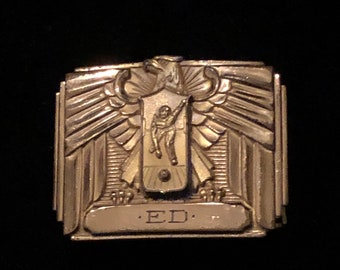 May include: A rectangular, silver-toned metal belt buckle with an Art Deco design. The buckle features an eagle with outstretched wings above a shield with a figure. The initials "ED" are engraved below.