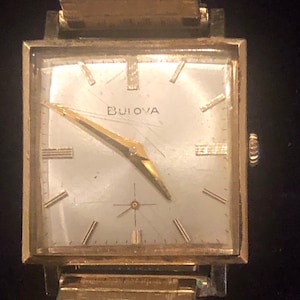 May include: A gold-toned Bulova wristwatch with a square face and a gold metal band. The watch face is white with black Roman numeral hour markers and black hands.