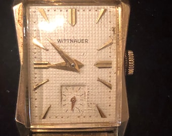 May include: A gold-toned Wittnauer wristwatch with a white face and black Roman numeral hour markers. The watch has a small second hand dial at the 6 o'clock position.