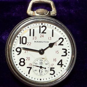 May include: A vintage Hamilton pocket watch with a white face, black numerals, and a smaller seconds dial. The watch has a silver-tone case and a loop for a chain. The word "HAMILTON" is printed on the face.