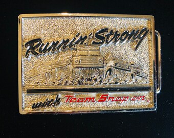 May include: A rectangular silver-colored belt buckle with the words "Runnin' Strong" in black script. Below the text are three silver-colored trucks. The words "with Team Snap-on" are in red and black.