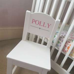 Pu&ograve; includere: Sedia in legno bianco con schienale arrotondato e il nome "POLLY" scritto in lettere rosa con un cuore grigio sopra il nome.