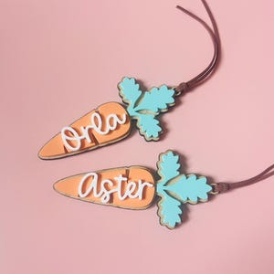 May include: Two wooden carrot ornaments with green tops and white lettering. The carrots are on a pink background. The top carrot says "Orla" and the bottom carrot says "Aster".