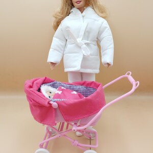 May include: A doll wearing a white quilted coat and white pants stands near a pink doll stroller. The stroller has a pink canopy and a baby doll wrapped in a pink blanket with white butterfly designs.