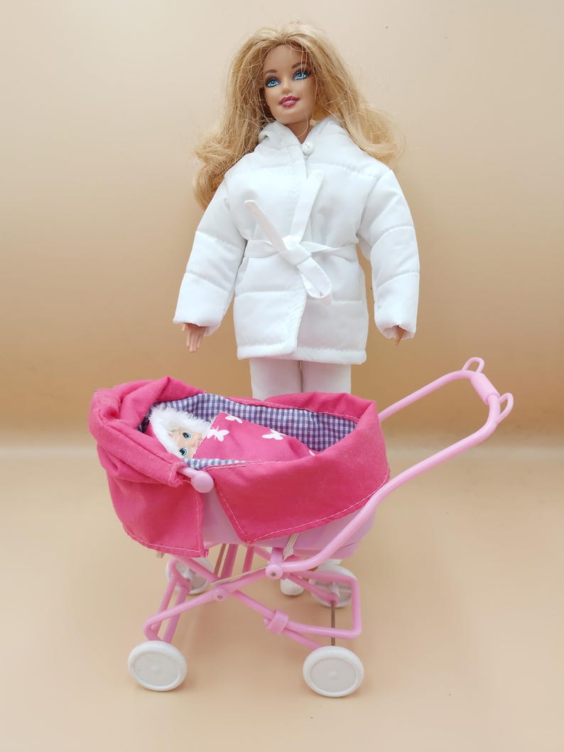 May include: A doll wearing a white quilted coat and white pants stands near a pink doll stroller. The stroller has a pink canopy and a baby doll wrapped in a pink blanket with white butterfly designs.
