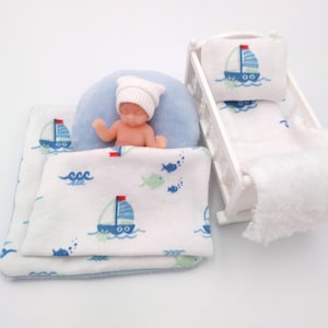 Könnte beinhalten: Eine Miniatur-Kinderzimmerszene mit einer Puppe mit weißer Strickmütze, die auf einem blauen Kissen liegt und mit einer weißen Decke mit Segelboot- und Fischmuster bedeckt ist. Ein weißes Kinderbett mit passender Bettwäsche ist ebenfalls vorhanden.