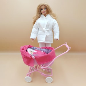 May include: A blonde doll wearing a white coat and white pants stands near a pink doll stroller. The stroller has a pink canopy and a baby doll wrapped in a pink blanket with white stars.